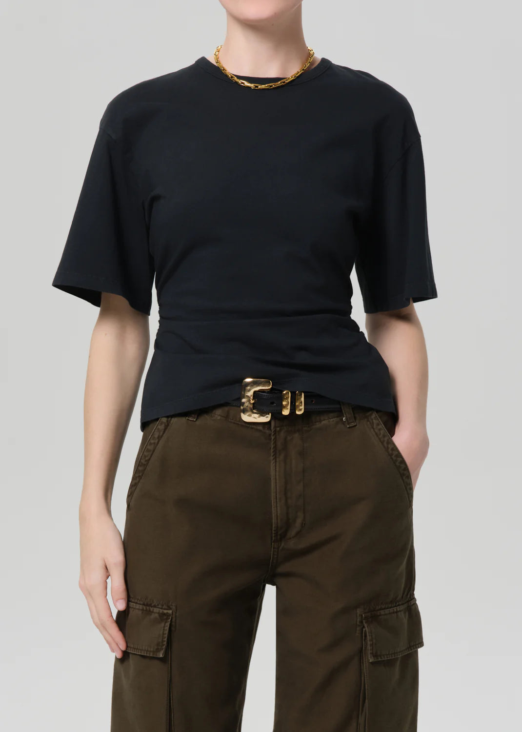 A person wears the Citizens Of Humanity/AGOLDE fabien tee, a black jersey cotton short-sleeve shirt with exaggerated sleeves, tucked into high-waisted dark olive cargo pants, accessorized with a gold belt and chain necklace against a light gray background.
