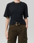A person wears the Citizens Of Humanity/AGOLDE fabien tee, a black jersey cotton short-sleeve shirt with exaggerated sleeves, tucked into high-waisted dark olive cargo pants, accessorized with a gold belt and chain necklace against a light gray background.