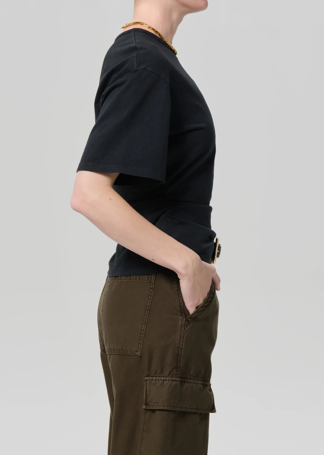 A person in profile against a plain background wears the Citizens Of Humanity/AGOLDE fabien tee—black jersey cotton, short-sleeve—paired with olive green cargo pants and a gold necklace, one hand casually in their pocket.