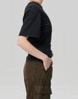 A person in profile against a plain background wears the Citizens Of Humanity/AGOLDE fabien tee—black jersey cotton, short-sleeve—paired with olive green cargo pants and a gold necklace, one hand casually in their pocket.