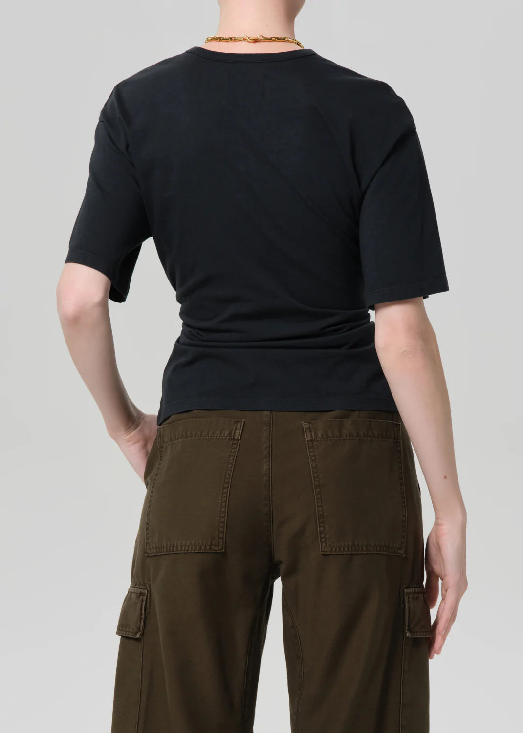 A person faces away from the camera, wearing the Citizens Of Humanity/AGOLDE fabien tee in black with exaggerated sleeves and olive green cargo pants, both set against a plain light gray background.