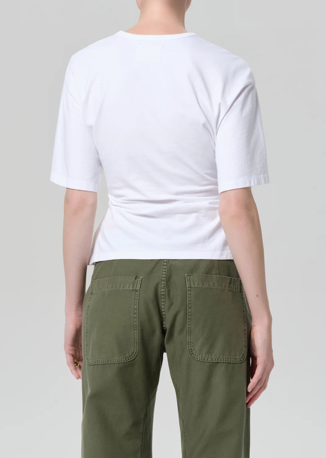 A person faces away from the camera, wearing the fabien tee by Citizens Of Humanity/AGOLDE—crafted from soft white jersey cotton—paired with green pants featuring large back pockets, set against a plain light gray backdrop.