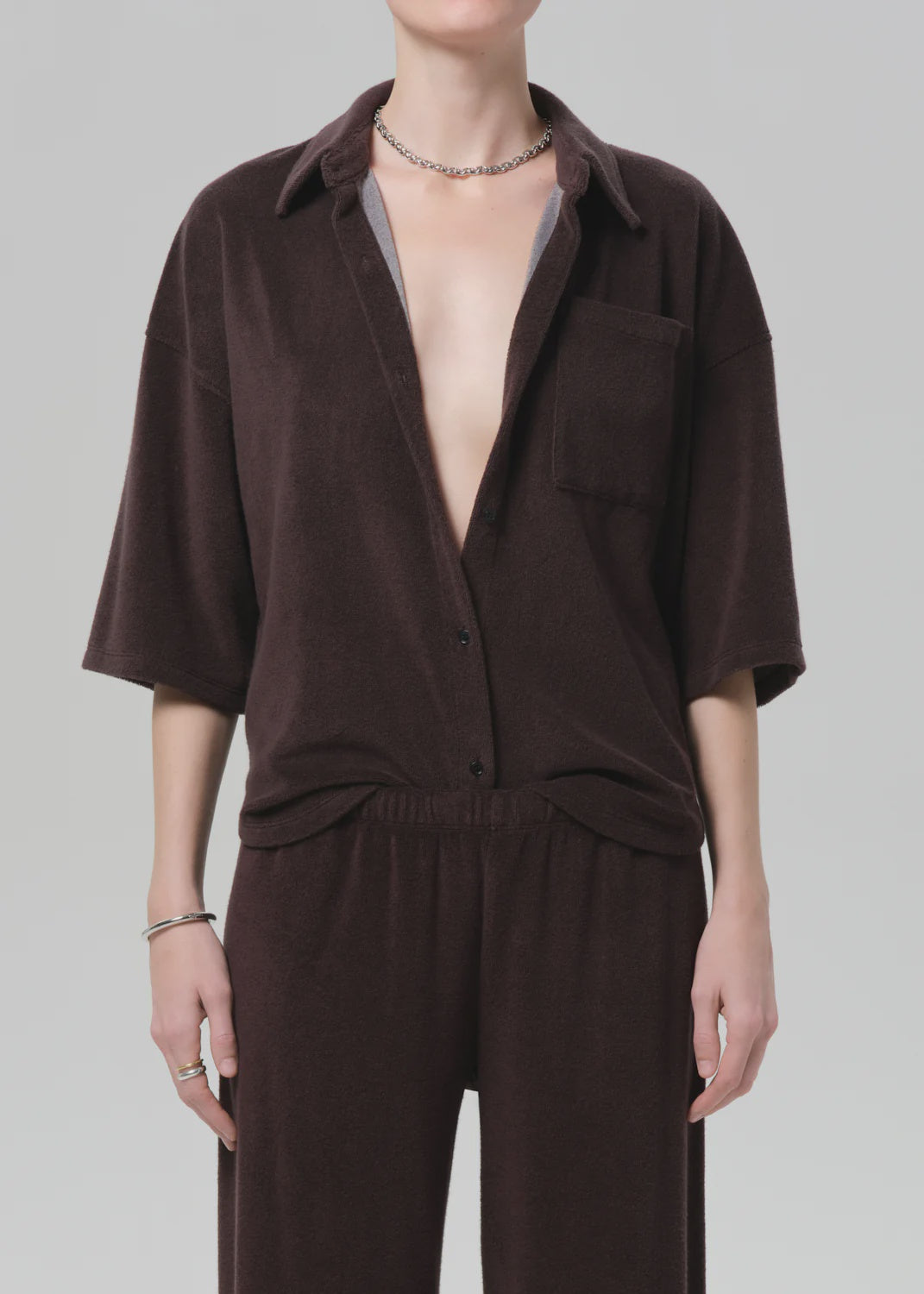 A person wears the Citizens Of Humanity/AGOLDE francis knit shirt in dark brown, styled partially unbuttoned with matching pants. They accessorize with a silver necklace and rings against a plain, light background.