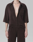 A person wears the Citizens Of Humanity/AGOLDE francis knit shirt in dark brown, styled partially unbuttoned with matching pants. They accessorize with a silver necklace and rings against a plain, light background.