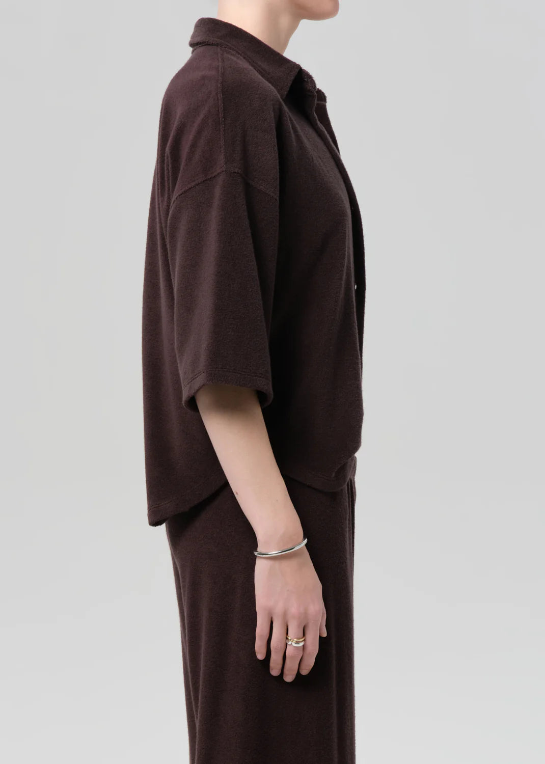 A person stands in profile against a plain background, wearing the Citizens Of Humanity/AGOLDE francis knit shirt in dark brown with short sleeves and a matching skirt. Their face is hidden, and they wear silver rings and a bracelet on their left hand.