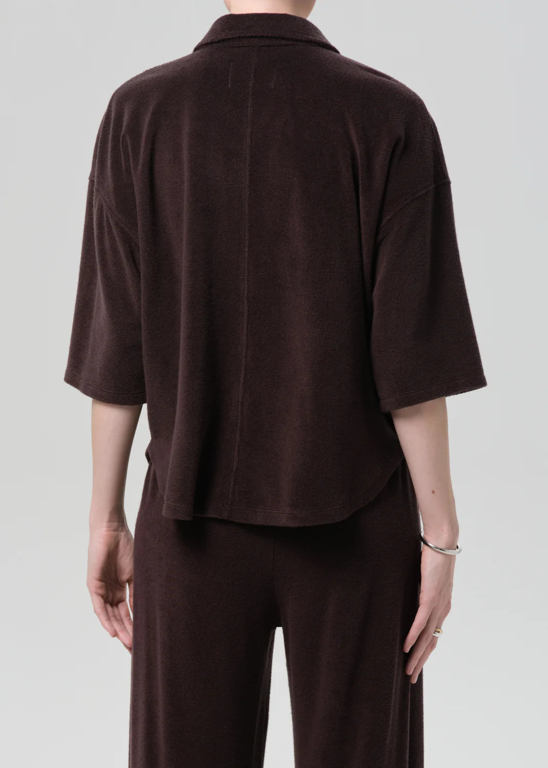 A person stands with their back to the camera in a loose-fitting, dark brown francis knit shirt by Citizens Of Humanity/AGOLDE and matching pants in a microterry cotton blend, short sleeve button front top contrasting a light gray background.