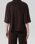 A person stands with their back to the camera in a loose-fitting, dark brown francis knit shirt by Citizens Of Humanity/AGOLDE and matching pants in a microterry cotton blend, short sleeve button front top contrasting a light gray background.