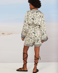 A person with short curly hair stands facing away in a cream long-sleeve top and Me369 Eleanor Bermuda Shorts featuring black doodle-style drawings. They wear black lace-up sandals on a sandy, desert-like landscape.