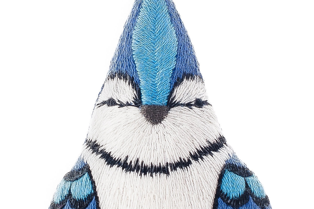 Close-up of a blue jay crafted with blue, black, and white thread from the Faire Blue Jay Embroidery Kit. The stitching creates a stylized, symmetrical Blue Jay Pattern on a white background.
