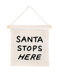 A Canvas Hang Sign by Faire, featuring a small beige design made from natural canvas, is suspended from a wooden dowel with "SANTA STOPS HERE" boldly screen printed in black letters.