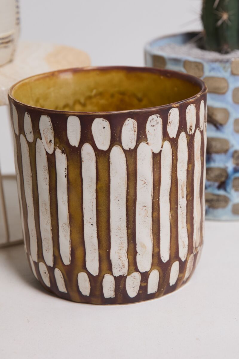 The Anselm Pot Brown by Accent Decor, featuring vertical white oval patterns on a brown background, sits on a light surface with a blue cactus pot in the background.