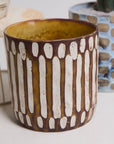 The Anselm Pot Brown by Accent Decor, featuring vertical white oval patterns on a brown background, sits on a light surface with a blue cactus pot in the background.