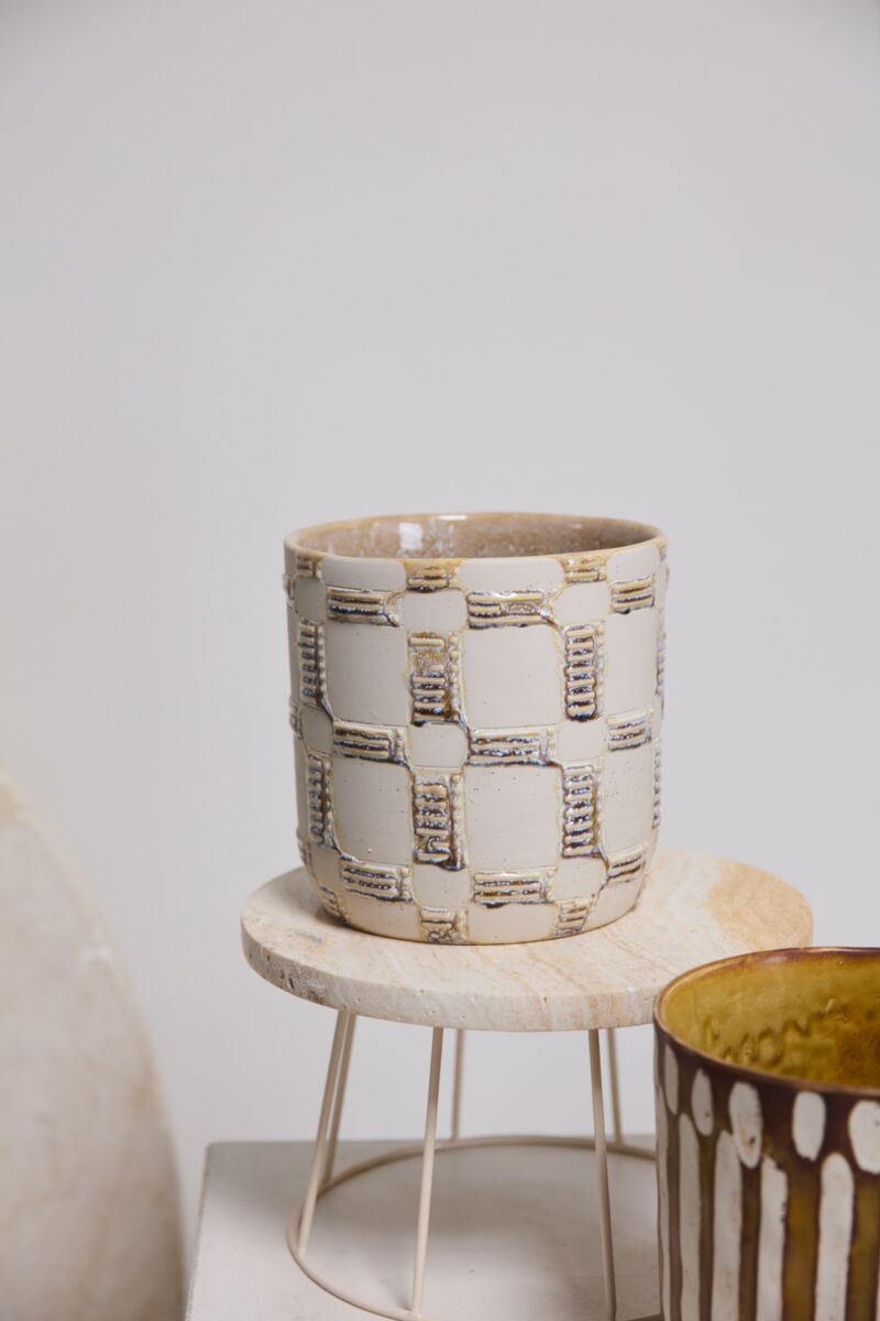 The Anselm Pot by Accent Decor features a hand-painted, textured geometric pattern in beige and white on a round wooden stand with metal legs. A partially visible artisanal planter displays a brown and white striped design beside it.