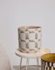 The Anselm Pot by Accent Decor features a hand-painted, textured geometric pattern in beige and white on a round wooden stand with metal legs. A partially visible artisanal planter displays a brown and white striped design beside it.