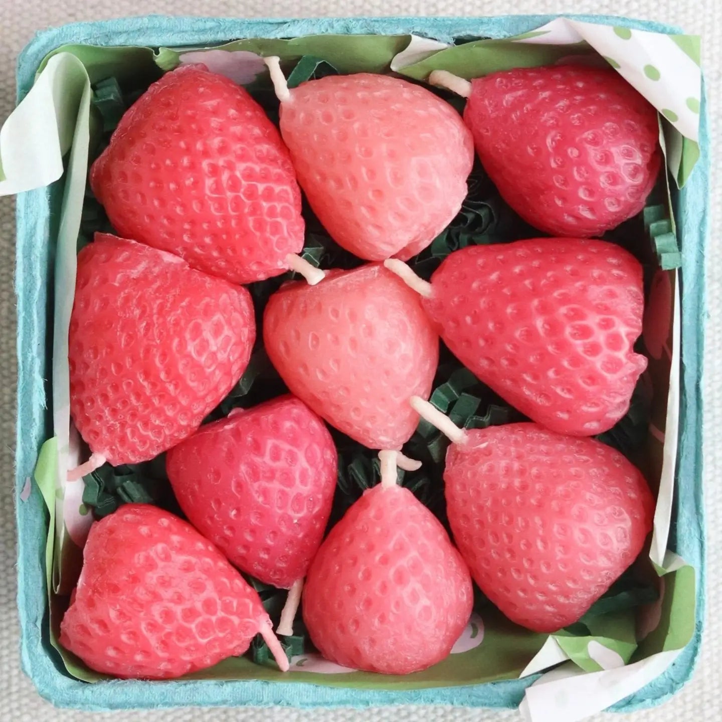 A box of twelve Faire Pink Strawberry Birthday Candles, shaped like strawberries with white wicks and arranged on green paper shreds. These eco-friendly candles add a charming touch to any celebration.