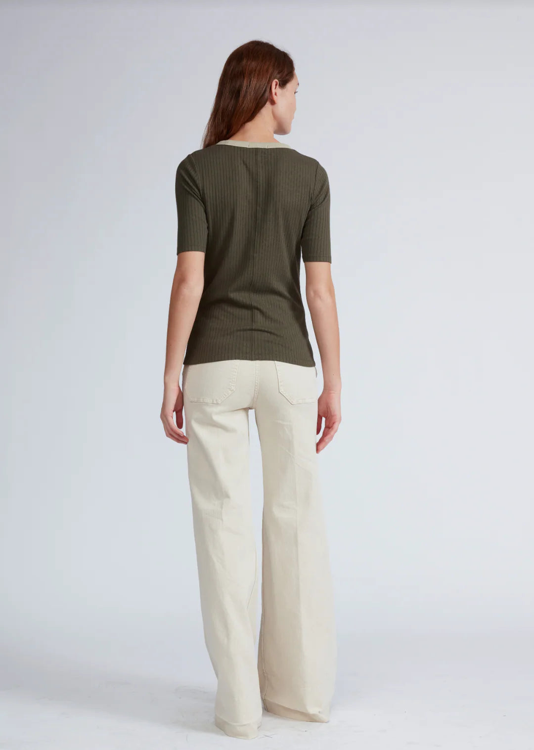 A woman with long brown hair, facing away, wears the knit contrast ringer tee by Rag & Bone in olive green and high-waisted cream wide-leg pants against a plain light gray background.