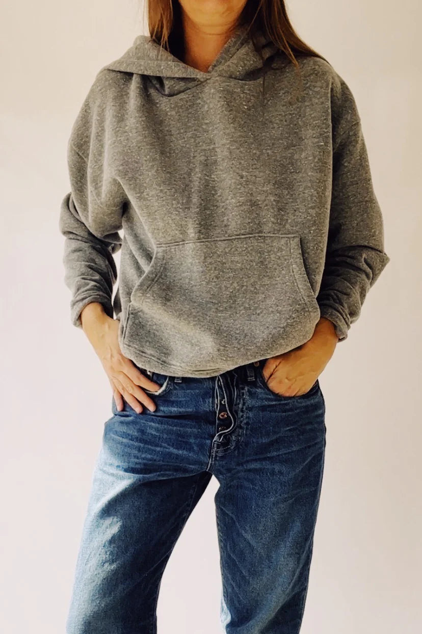 A person wearing &quot;The Hoodie&quot; by Hey Gang in a boxy fit and blue jeans stands with hands in pockets against a neutral background. This gray hoodie, made from 100% cotton, provides comfort and style with its unisex sizing.