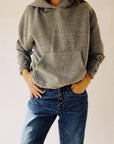 A person wearing "The Hoodie" by Hey Gang in a boxy fit and blue jeans stands with hands in pockets against a neutral background. This gray hoodie, made from 100% cotton, provides comfort and style with its unisex sizing.