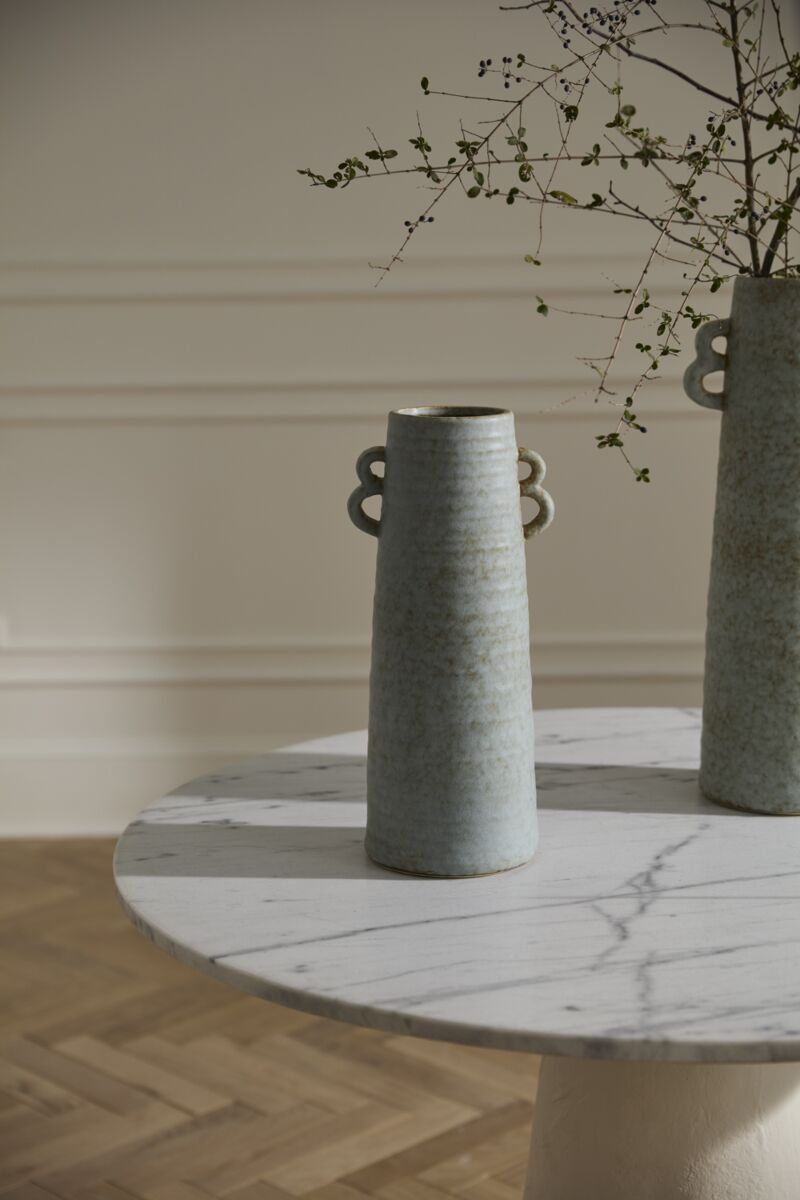 The Bowen Vase M by Accent Decor, a tall ceramic piece with handles and a soft blue glaze, sits on a round white marble table. Sparse leafy branches are arranged inside, with a cream paneled wall and wooden floor in the background.