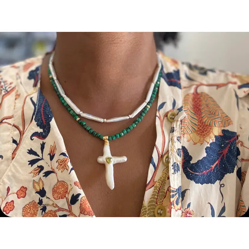 A person wearing a floral print shirt and two necklaces: one with malachite cubes and white beads, the other—the Carmelia Necklace by Faire—with green beads and a large peridot cross pearl pendant.