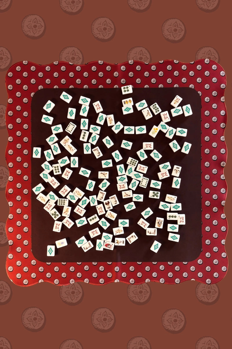 Wild West Mahjong SET by Faire: Tiles scattered on a dark square mat with a red scalloped border, resting on a brown patterned background—capturing the classic Western Mahjong style. Includes tiles, 4 racks, bag, and mat.