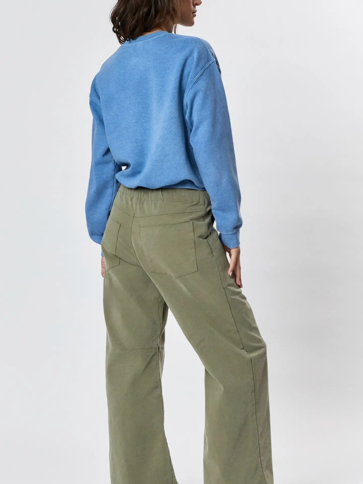Person standing with their back to the camera, wearing a blue long-sleeve top and Faire's Gwen Boyfriend Wide Leg Pant in olive green organic cotton, set against a plain light background.