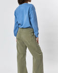 Person standing with their back to the camera, wearing a blue long-sleeve top and Faire's Gwen Boyfriend Wide Leg Pant in olive green organic cotton, set against a plain light background.