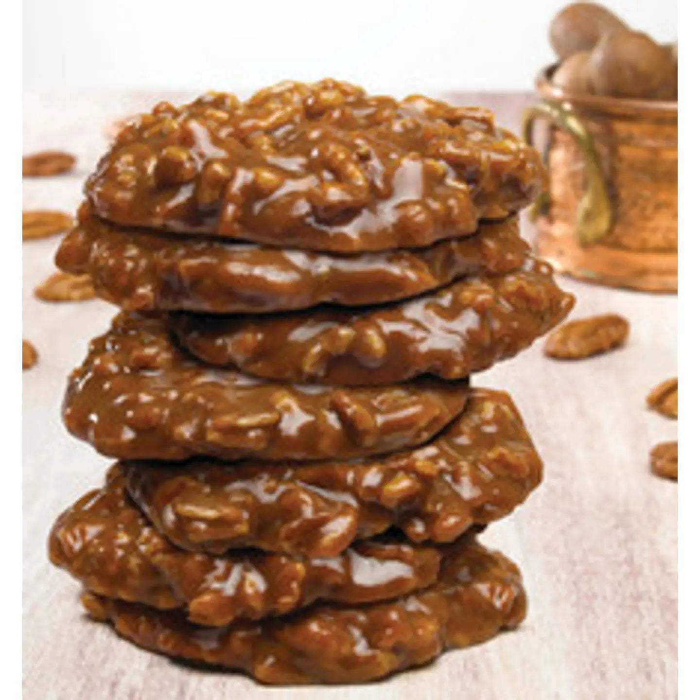 A stack of Faire LC Praline Big Chews 2.5oz cookies sits on a light wooden surface, surrounded by pecan pieces, with a copper pot softly blurred in the background.