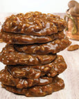 A stack of Faire LC Praline Big Chews 2.5oz cookies sits on a light wooden surface, surrounded by pecan pieces, with a copper pot softly blurred in the background.