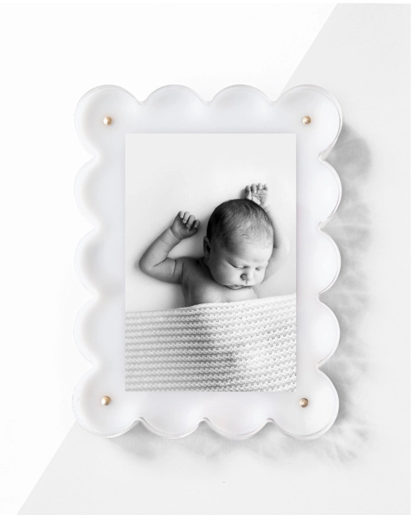 A monochrome photo of a sleeping baby under a textured blanket is elegantly framed in Faire&#39;s Acrylic Frame, which features a decorative scalloped white design with gold accents that casts a soft shadow on the white surface behind it.