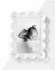 A monochrome photo of a sleeping baby under a textured blanket is elegantly framed in Faire's Acrylic Frame, which features a decorative scalloped white design with gold accents that casts a soft shadow on the white surface behind it.