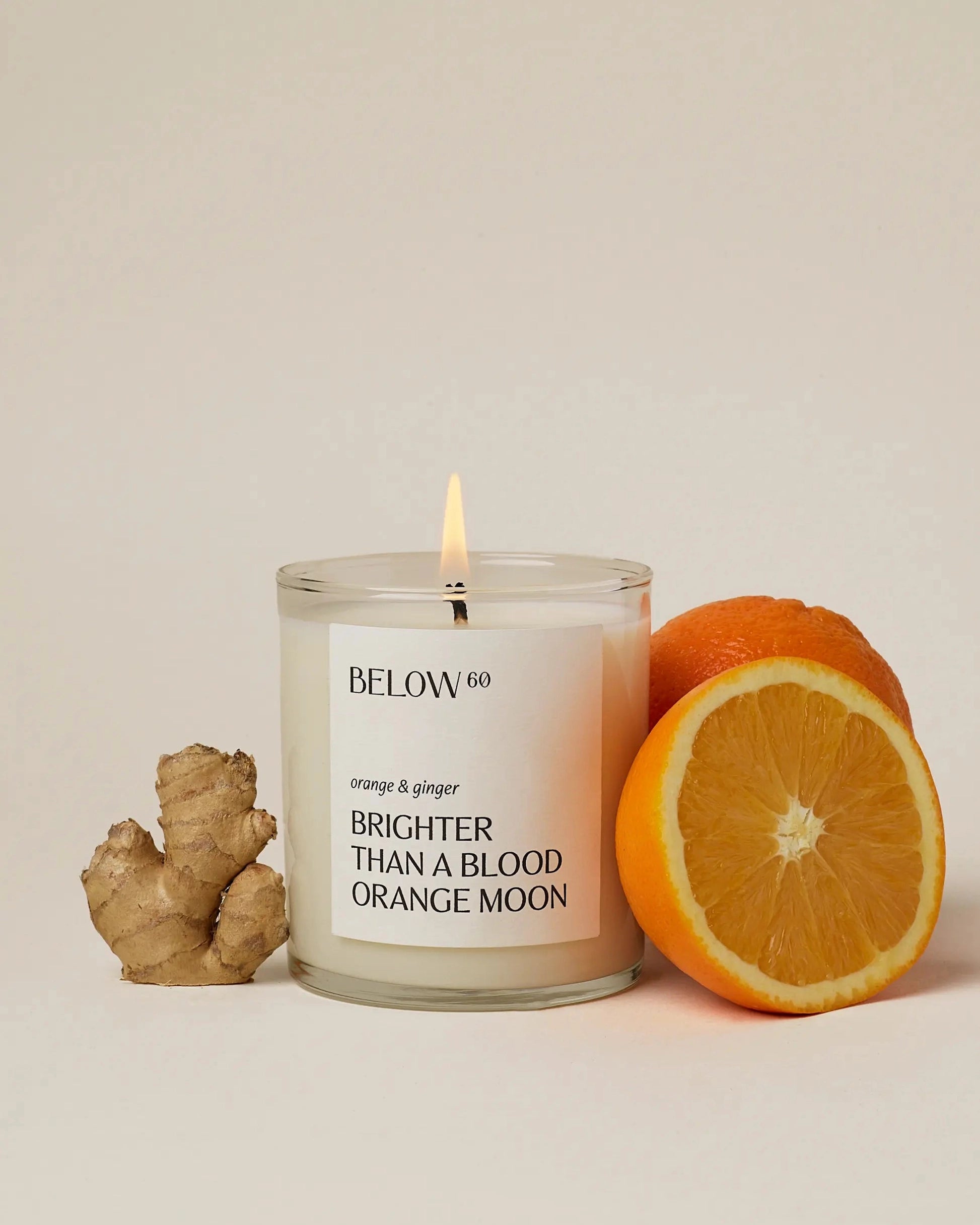 A lit Below 60 Candle by Faire with orange & ginger scent sits next to fresh ginger root and an orange, one half sliced open, on a neutral background.