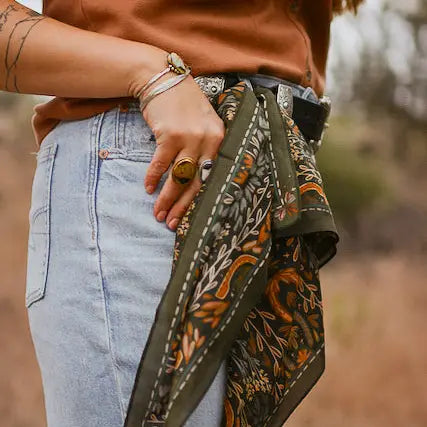 A person outdoors wears light blue jeans, a brown shirt, multiple rings and bracelets, with the Faire Wild Rag Silk Scarf in green tied to their belt loop as a chic cowgirl accessory.