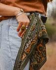 A person outdoors wears light blue jeans, a brown shirt, multiple rings and bracelets, with the Faire Wild Rag Silk Scarf in green tied to their belt loop as a chic cowgirl accessory.