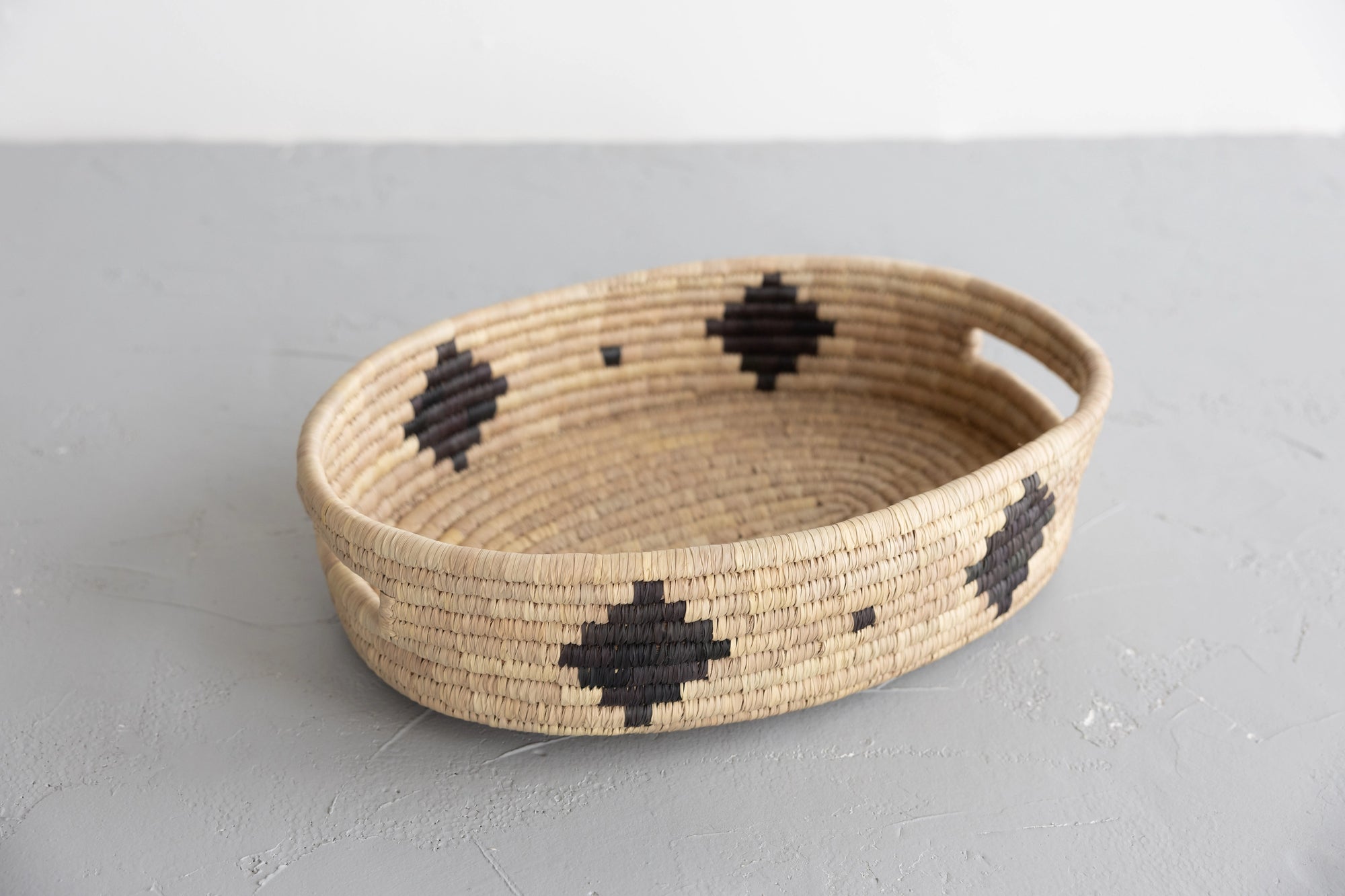 The Faire Palm Leaf Oval Basket is a shallow, handwoven tray with two handles and bold black geometric patterns, crafted by fair trade artisans. It rests on a light gray textured surface.