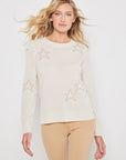 A person with wavy, blonde hair elegantly poses against a white background, wearing Lisa Todd's luxury cashmere "Star Dust" sweater adorned with contrast intarsia stars and beige pants.