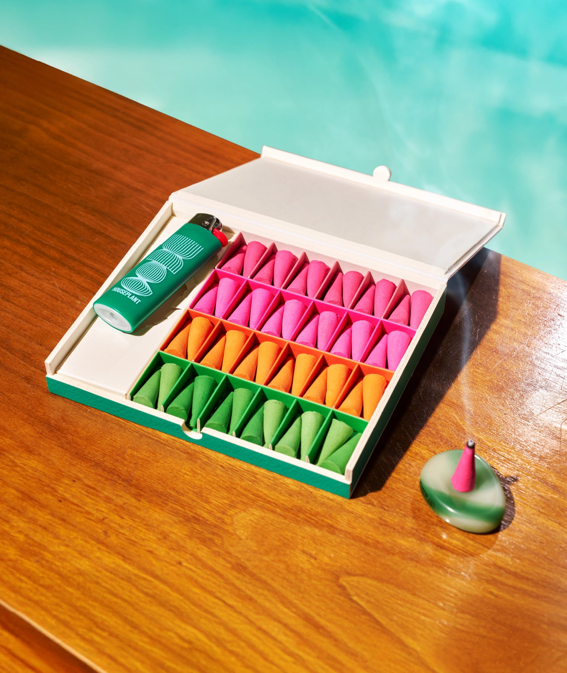 Houseplant by Ripple Incense Droplets from Faire, with a green lighter and one burning on a ceramic holder, add vibrant home fragrance to your poolside table.