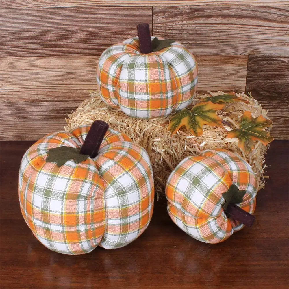 Three Faire Pumpkin decorations in orange, green, and white plaid rest on a wooden surface—two on the table and one on a hay bale with autumn leaves—adding cozy farmhouse fall charm against a wood panel backdrop.