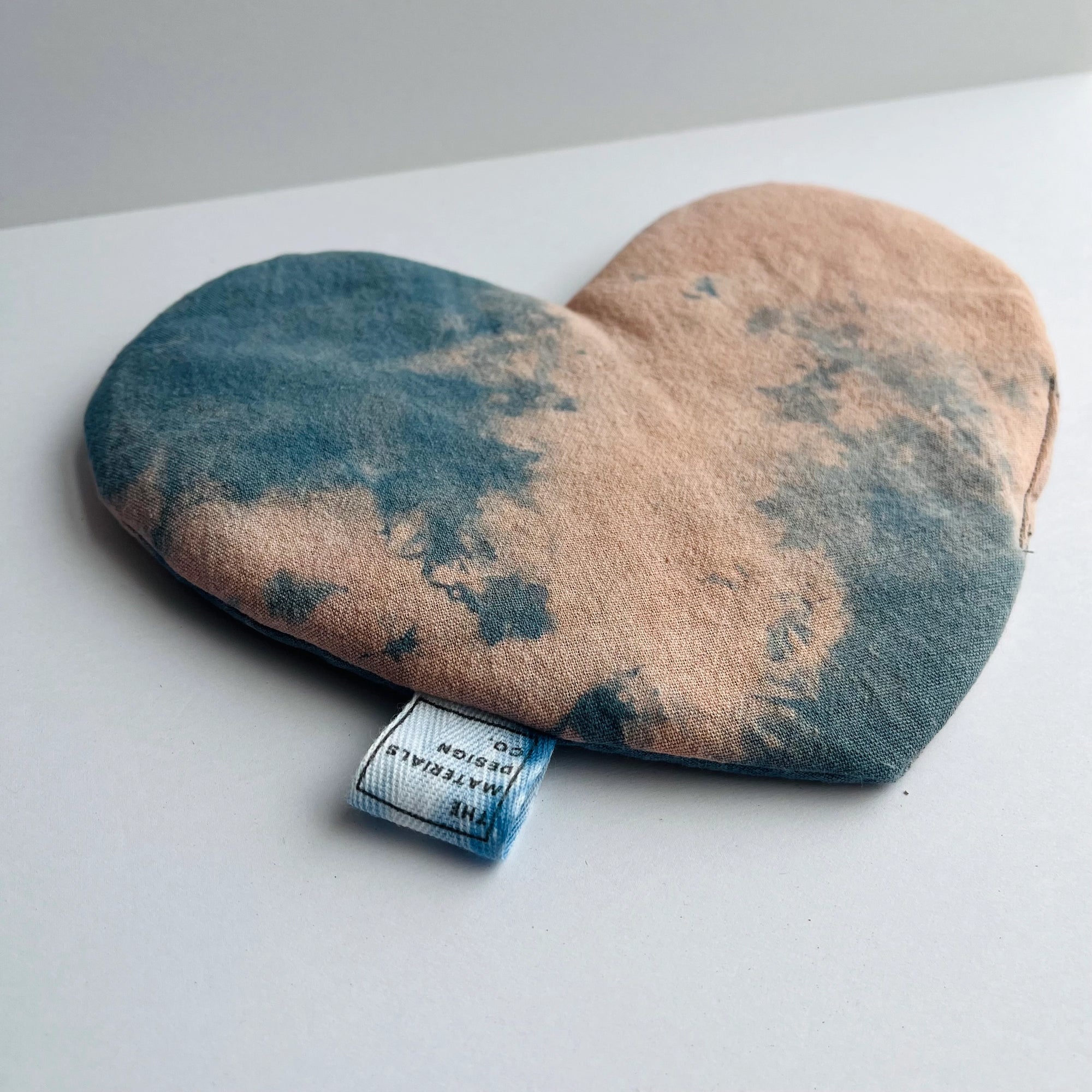 The Faire Heart Eye Pillow, featuring a blue and peach tie-dye cotton design, rests on a white surface. This heart-shaped pouch is perfect for anxiety relief and includes a small tag at the bottom edge.