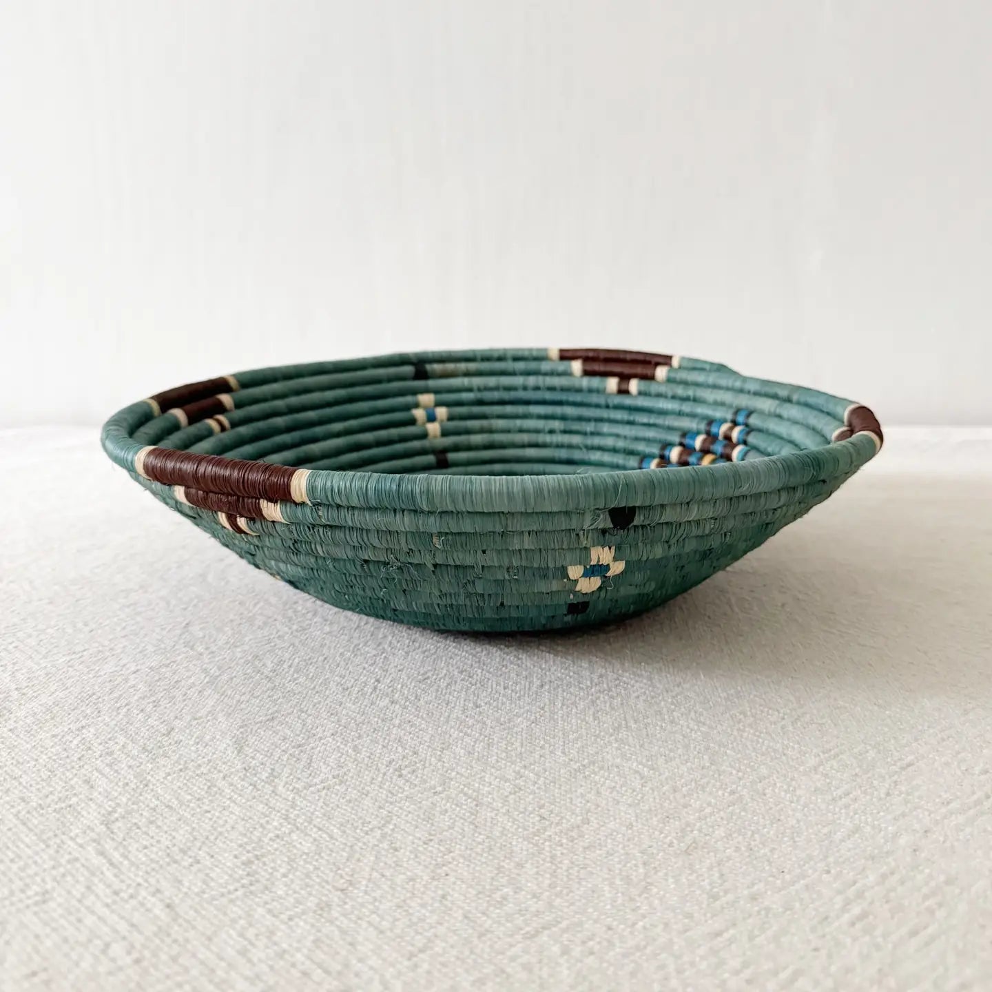 The Birava Large Bowl by Faire is a shallow, round woven basket in teal, brown, cream, and blue—ideal as handmade decor or Rwanda wall art. Shown on a light textured surface with a plain white background.