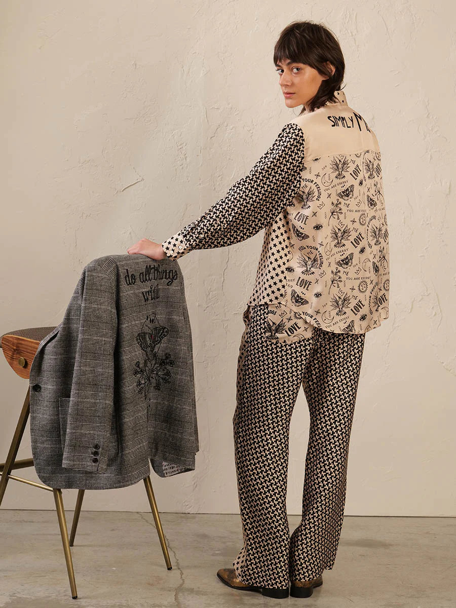 A person in Me369’s Marilyn Trousers stands by a stool, glancing over their shoulder and holding a gray jacket with a black embroidered design and the words "do all things with love.