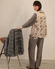 A person in Me369’s Marilyn Trousers stands by a stool, glancing over their shoulder and holding a gray jacket with a black embroidered design and the words "do all things with love.