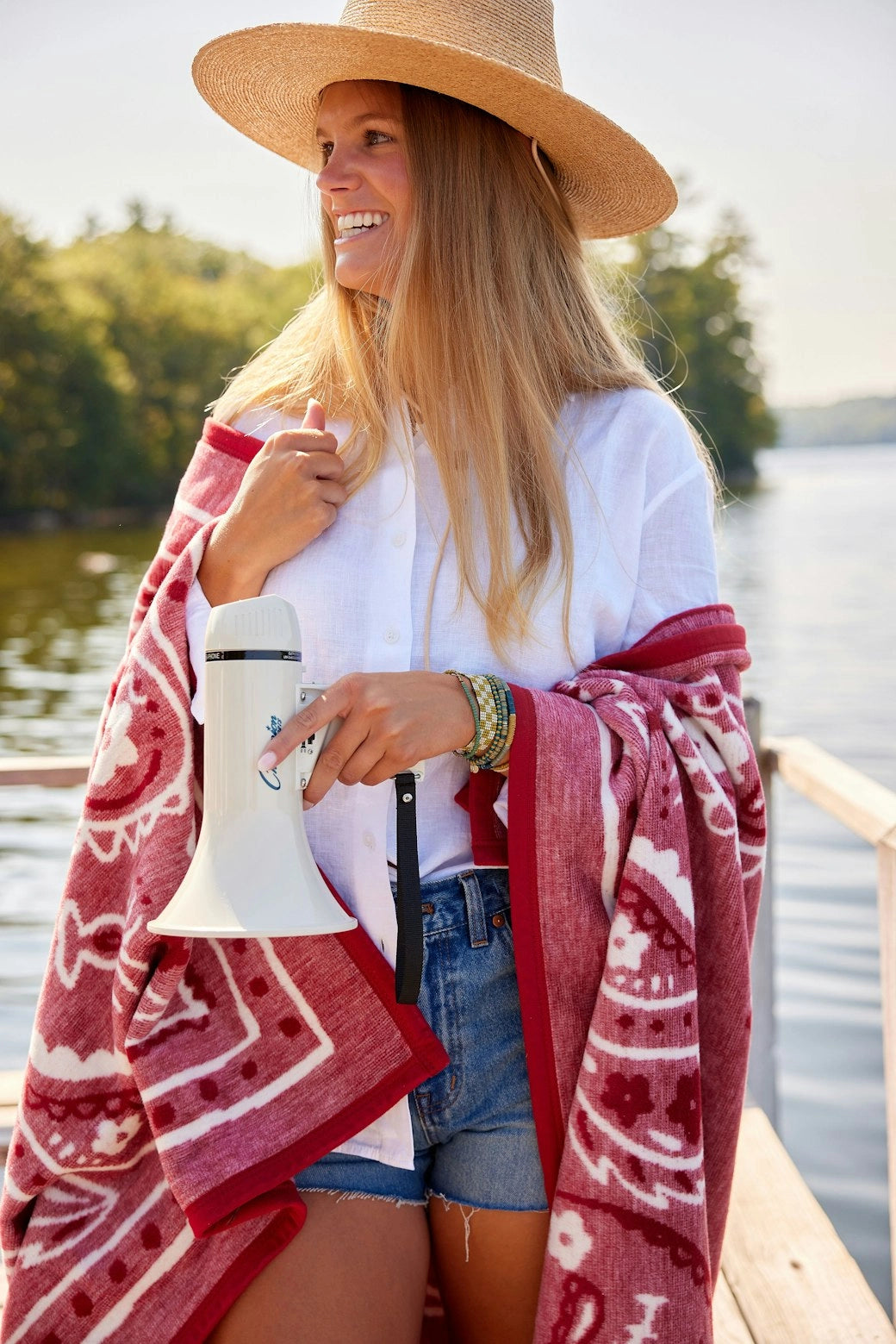 A woman in a straw hat, white shirt, and denim shorts stands on a dock wrapped in the Faire Vintage Bandana Blanket, megaphone in hand. Trees and water create an American spirit backdrop.