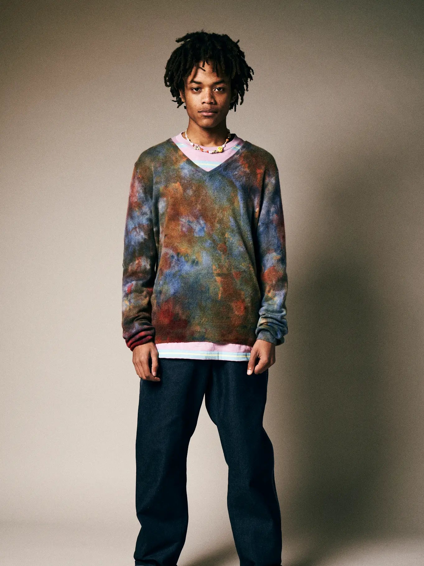 A person with medium-length locs models the Riverside Tool & Dye Cashmere V-neck sweater—loose, multicolored, and unisex—over a white shirt and dark blue pants, with a beaded necklace against a neutral beige background.