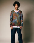 A person with medium-length locs models the Riverside Tool & Dye Cashmere V-neck sweater—loose, multicolored, and unisex—over a white shirt and dark blue pants, with a beaded necklace against a neutral beige background.