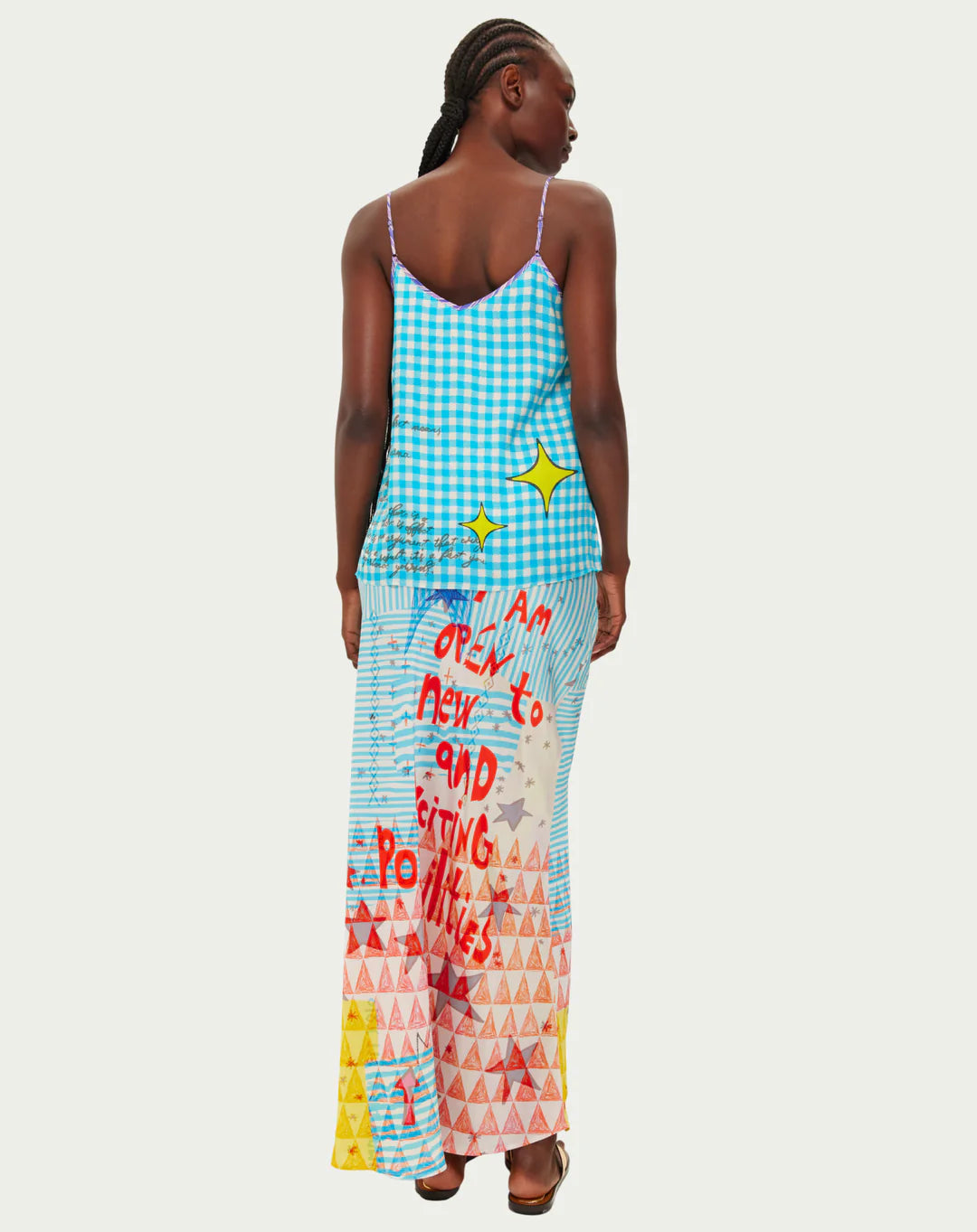 A woman faces away, wearing the Me369 Angelique Camisole: a turquoise gingham cami top paired with a patterned skirt featuring red "I am open to new and exciting possibilities" text and yellow, red, blue geometric shapes.