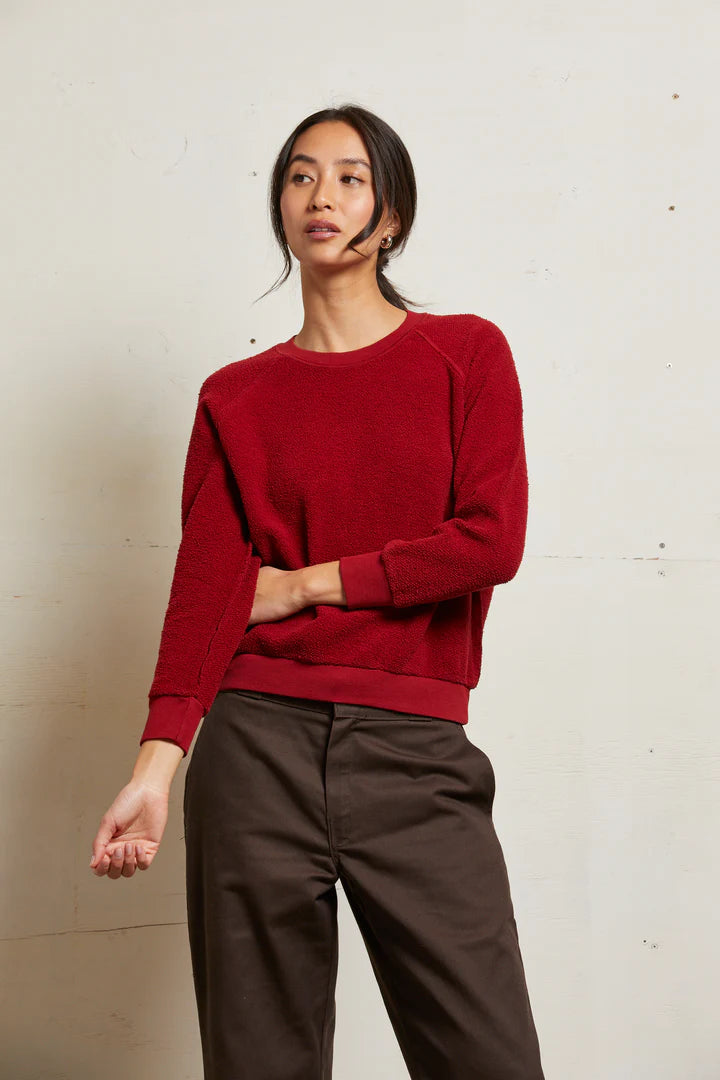 A woman with long dark hair pulled back stands against a light wall, wearing Perfectwhitetee's Ziggy Reverse LS Sweatshirt in a shrunken fit and brown pants, resting one hand on her opposite arm and gazing slightly to the side.