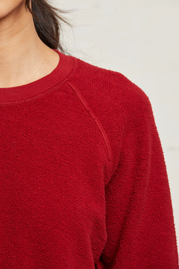 Close-up of a person wearing the Perfectwhitetee Ziggy Reverse LS Sweatshirt in textured red, showcasing its shrunken fit and crew neckline. The neutral background highlights the sweater&#39;s details around the shoulder and upper chest.