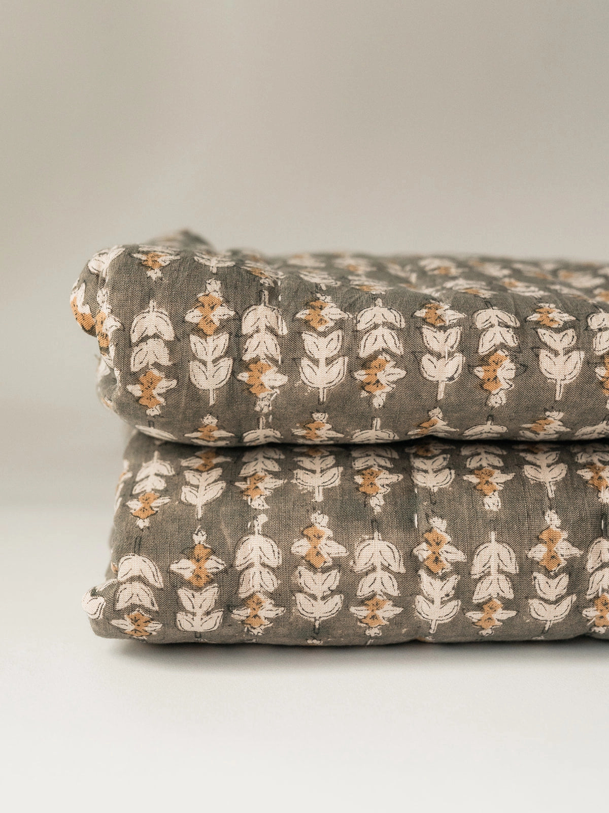 A close-up of two folded Faire Blossom Block Print Quilts with a white and orange floral pattern, stacked against a plain, light background.