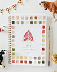 The Faire Watercolor Workbook is a beginner-friendly, spiral-bound book with a red barn cover featuring paint swatches. Nearby are a paintbrush, tube of paint, toy horse, cow, wheat stalks, and checkered bunting.